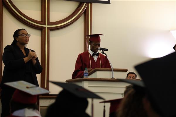 A graduate from the Alabama School for the Deaf speaks into a microphone from behind a podium. He is wearing his maroon Cap and Gown and a yellow cord. 